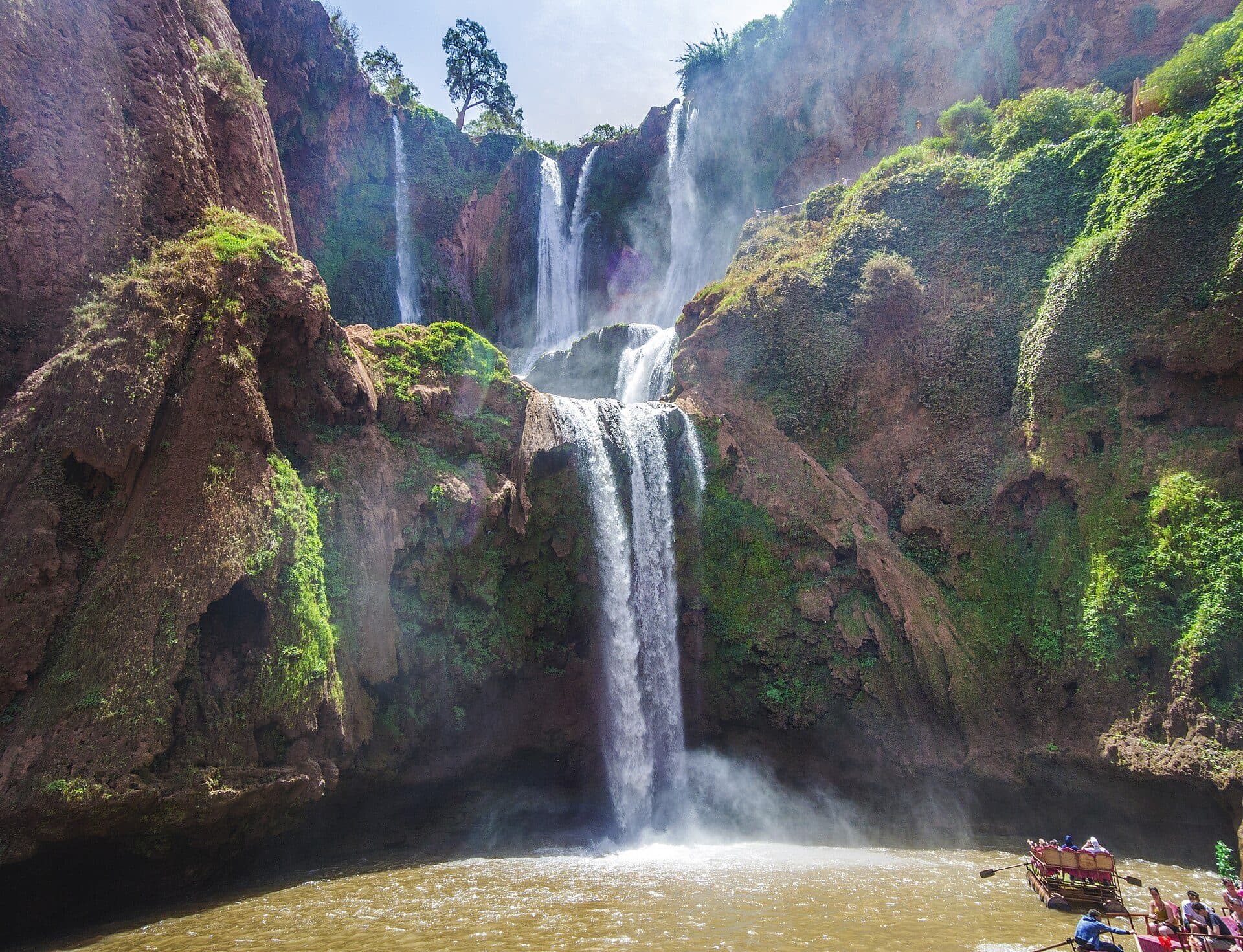 The Ouzoud waterfalls drop in three tiers down red, overgrown cliffs into a brown pool, with a raft full of people in the lower right corner
