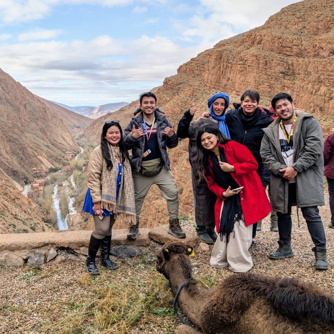 A group of travelers smiling at the camera above the Dadès Gorge, with the river far below and a resting camel in the foreground