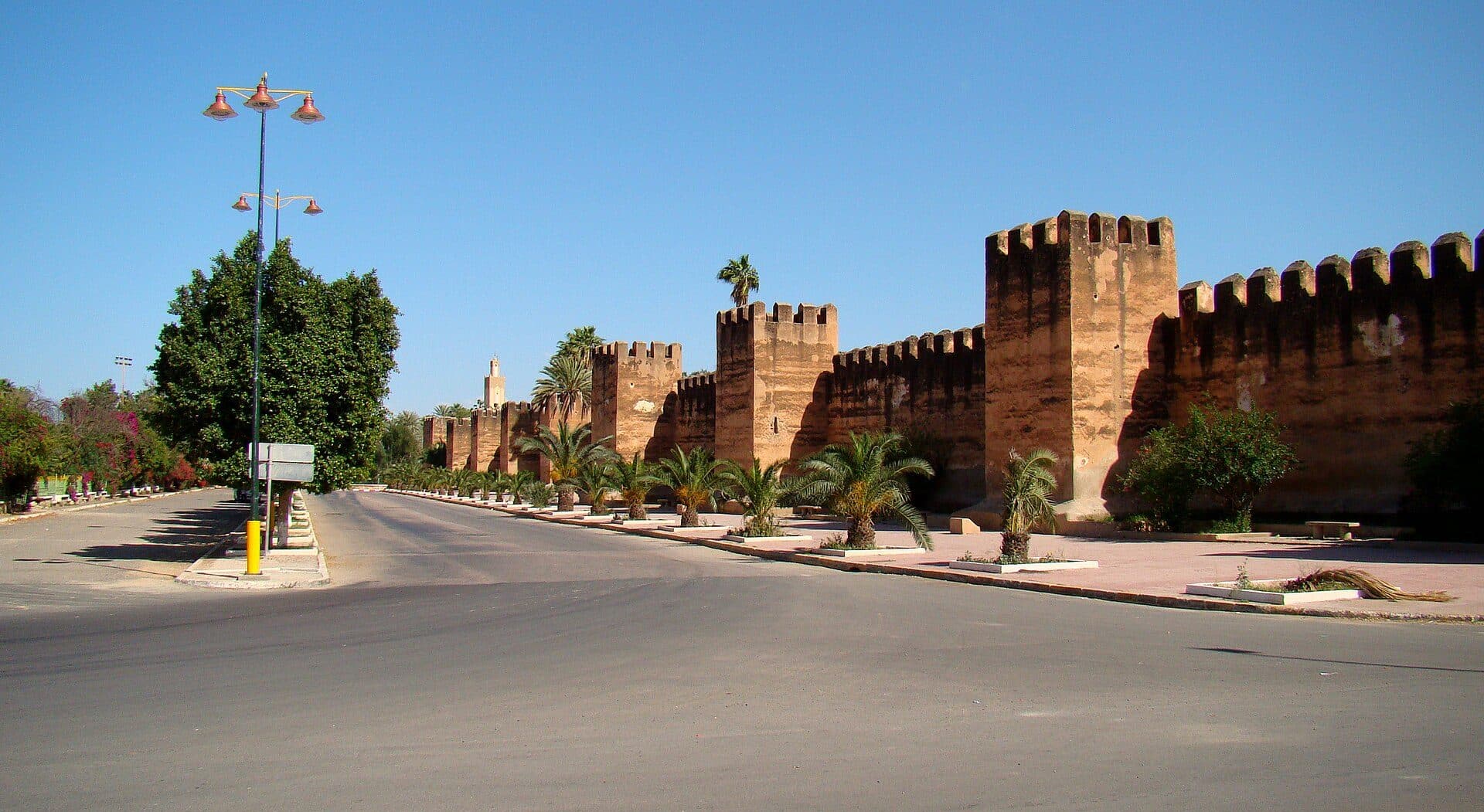 The mud-brick city wall of Taroudant with battlements and square towers, with palm trees and a wide road in front