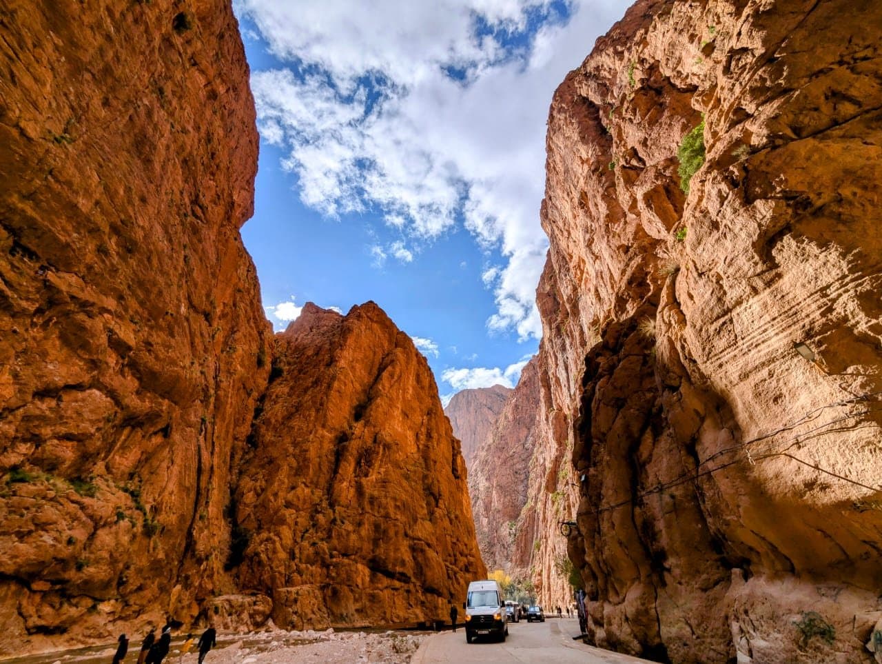 A van drives between the tall red rock walls of the Todra Gorge, with clouds in the narrow strip of sky above